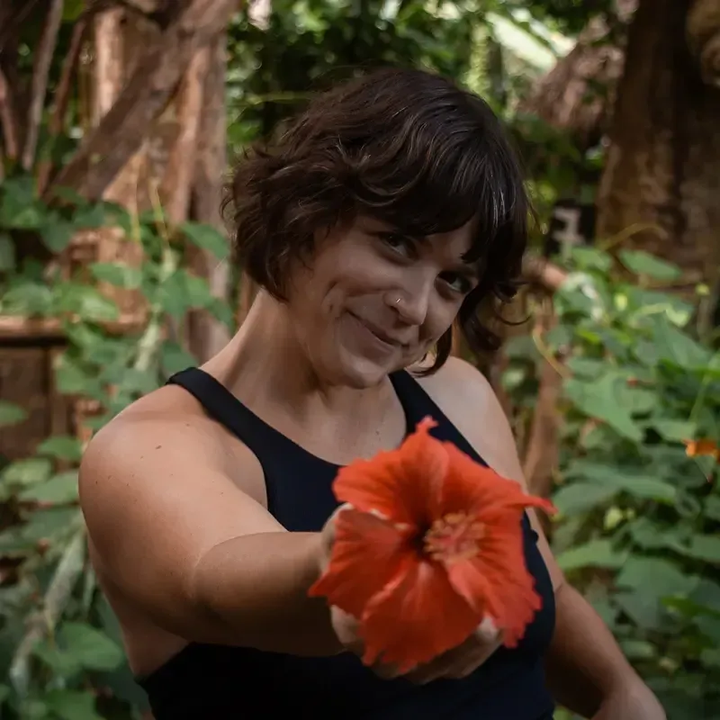 A smiling person holding out a bright orange hibiscus flower toward the camera in a lush, green garden setting.