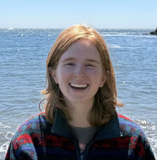 Smiling person in a red plaid jacket standing by the ocean on a sunny day