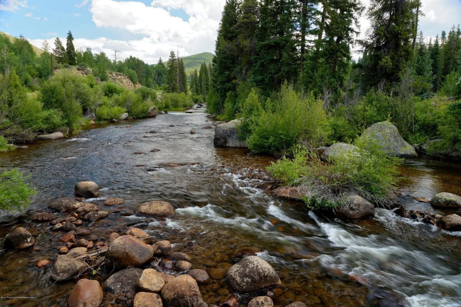 A mountain stream flows over rocky beds, surrounded by dense green trees and bushes under a partly cloudy sky.