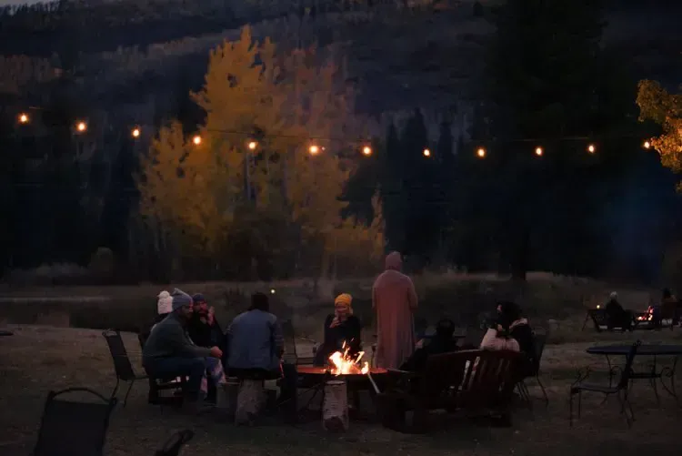 A group of people gathers around a glowing fire pit at dusk, under a string of warm fairy lights in a wooded setting.