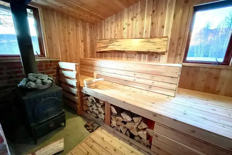 A wooden sauna interior featuring a stone-topped wood stove, tiered benches, and a stack of firewood underneath.