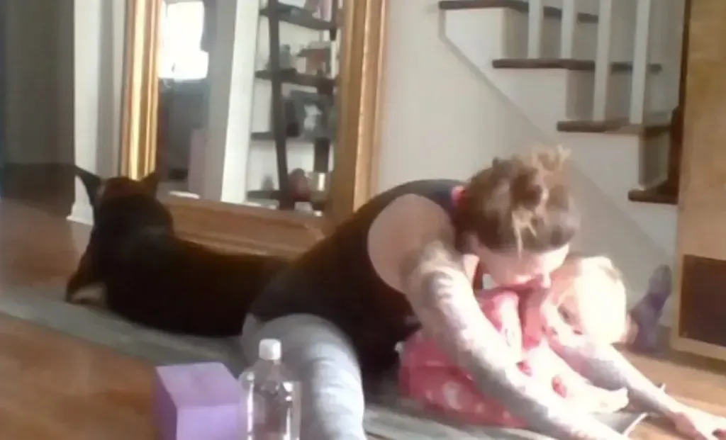 A person does floor stretches with a child and a dark-furred dog on a mat in a home setting.