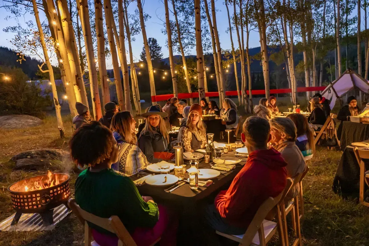 A group of people dining at long tables in a forest at twilight, illuminated by string lights and a nearby fire pit.