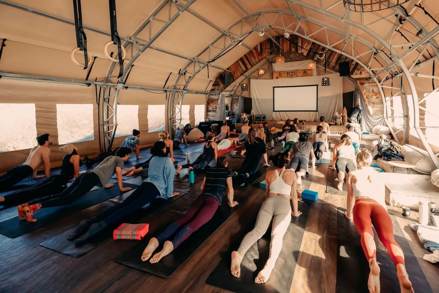 A group of people performing yoga on mats inside a large, open-air tent with a projection screen at the front.