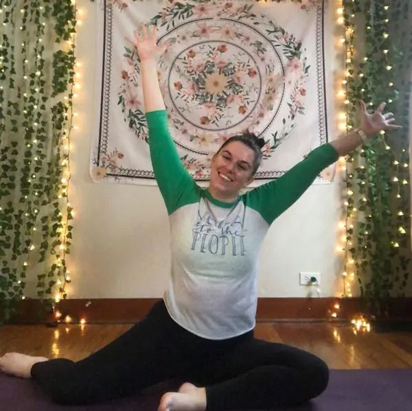 A smiling person in a green and white shirt sits on a yoga mat with arms raised in a V-shape, framed by fairy lights.
