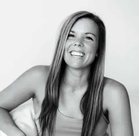 A black-and-white portrait of a smiling person with long, straight hair, wearing a tank top against a plain background.