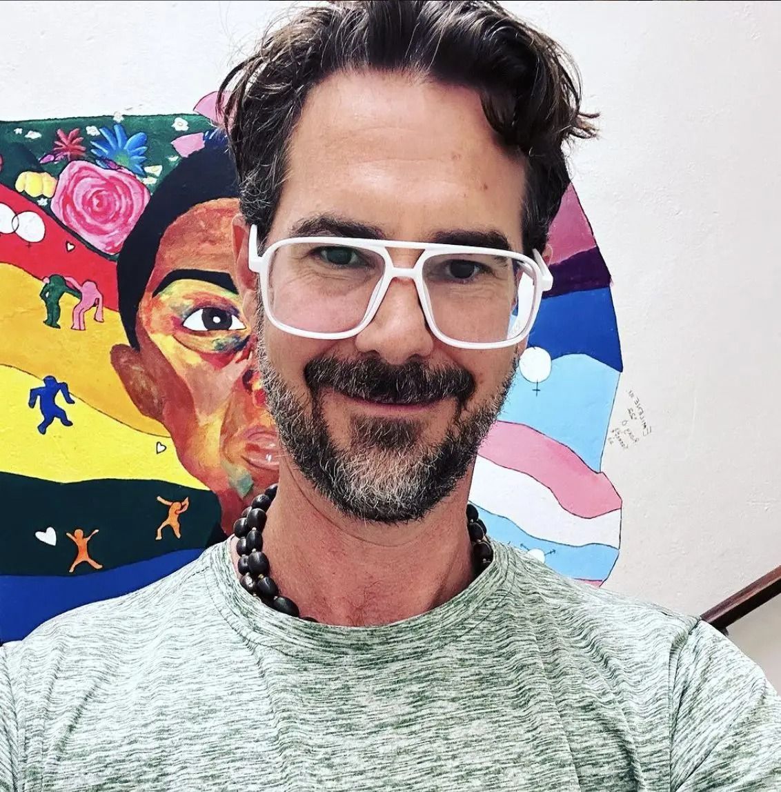 A person with a beard and white-framed glasses smiles in front of a colorful, abstract painting.