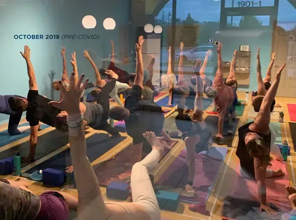 A yoga class performing side planks with raised arms in a studio with purple and blue mats on a wooden floor.