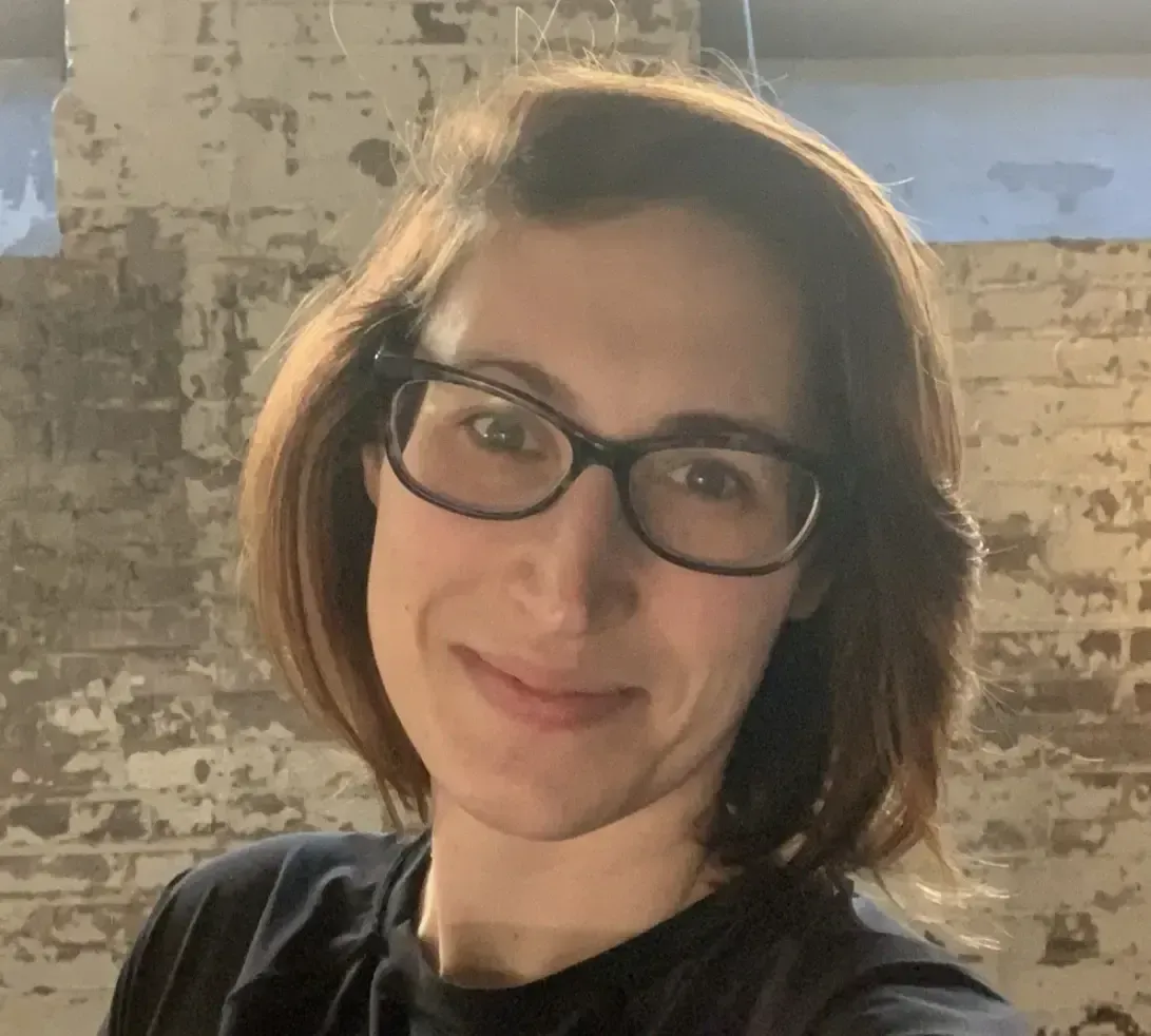 A person with shoulder-length brown hair and glasses smiling against a textured, light-colored brick wall.