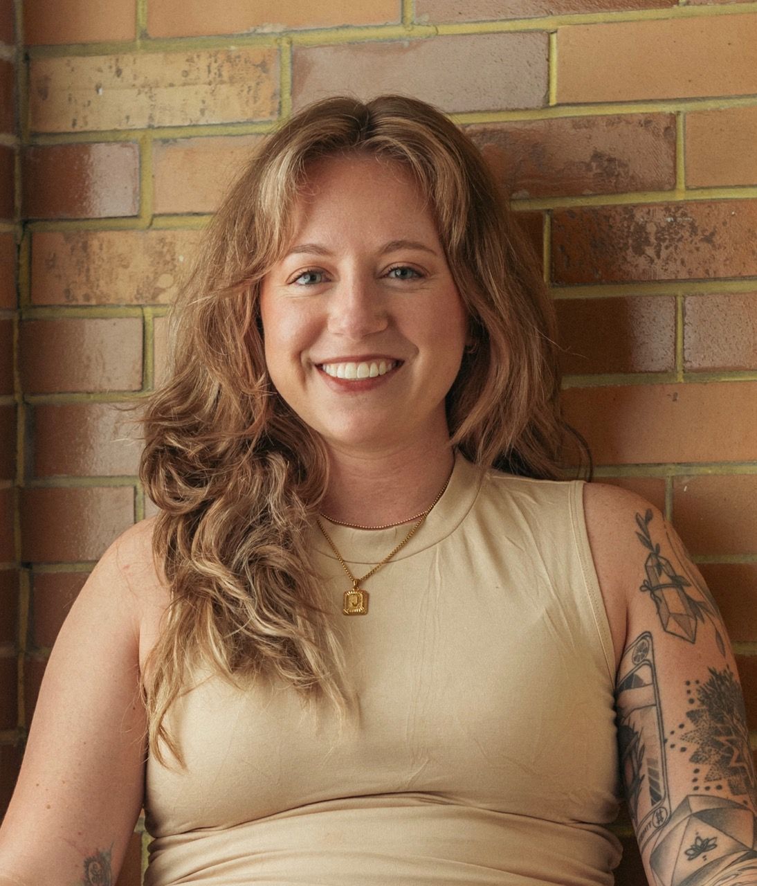 Smiling person in a beige sleeveless top, seated against a brick wall with visible tattoos.