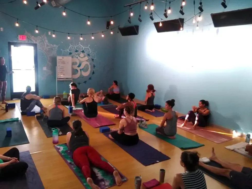 A yoga class in a blue studio with string lights, where participants on mats are following a teacher at the front.