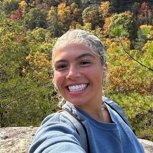 A person with light-colored hair and a blue sweatshirt smiles broadly in front of a background of autumn trees.