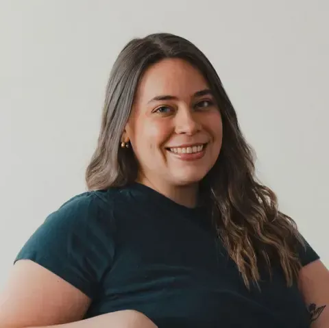 A person with long brown hair smiling against a plain, light-gray background while wearing a dark-colored shirt.