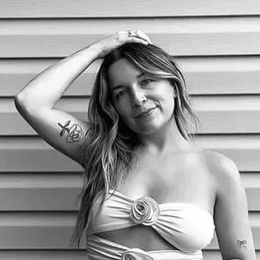 A person with wavy hair poses with their hand behind their head, wearing a strapless top, against a paneled background.