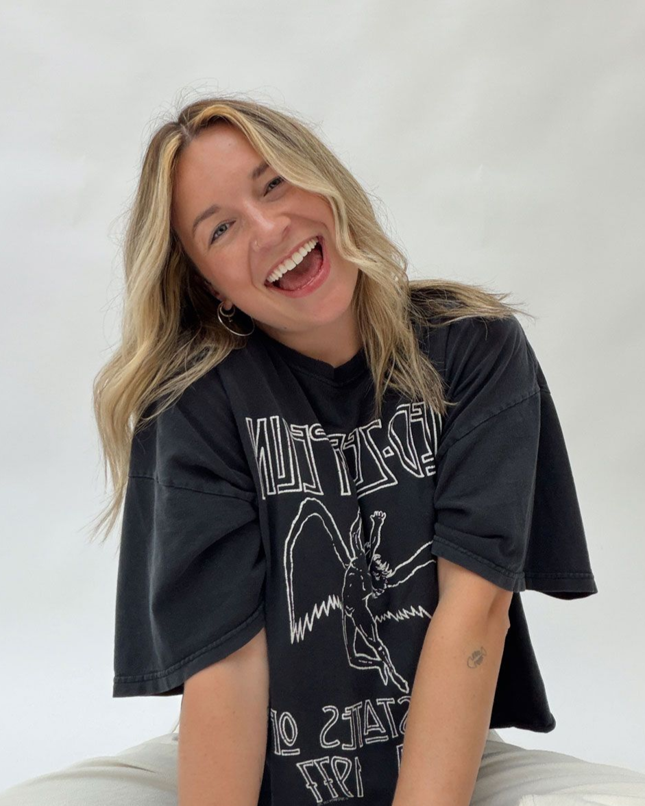 A person with wavy hair poses with their hand behind their head, wearing a strapless top, against a paneled background.