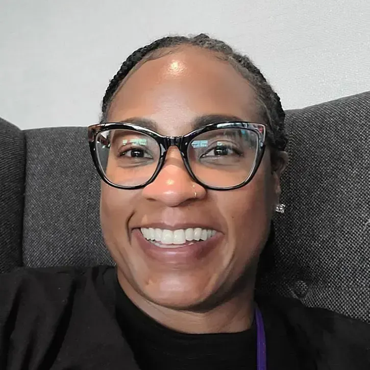A smiling person wearing black-rimmed cat-eye glasses, sitting in a gray fabric chair.