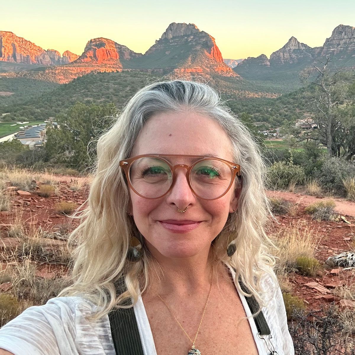 Smiling woman in glasses taking a selfie with red rock mountains at sunset behind her
