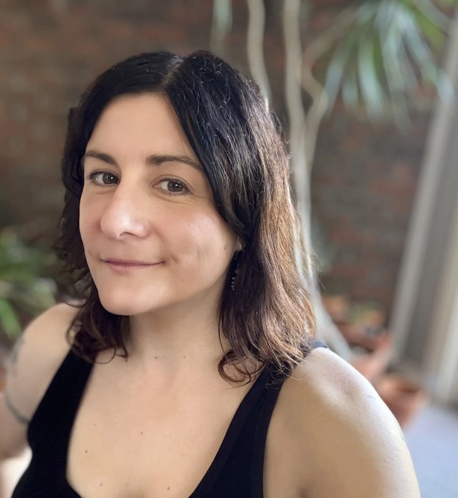 A person with shoulder-length dark hair and a light smile, wearing a black tank top in an indoor setting.