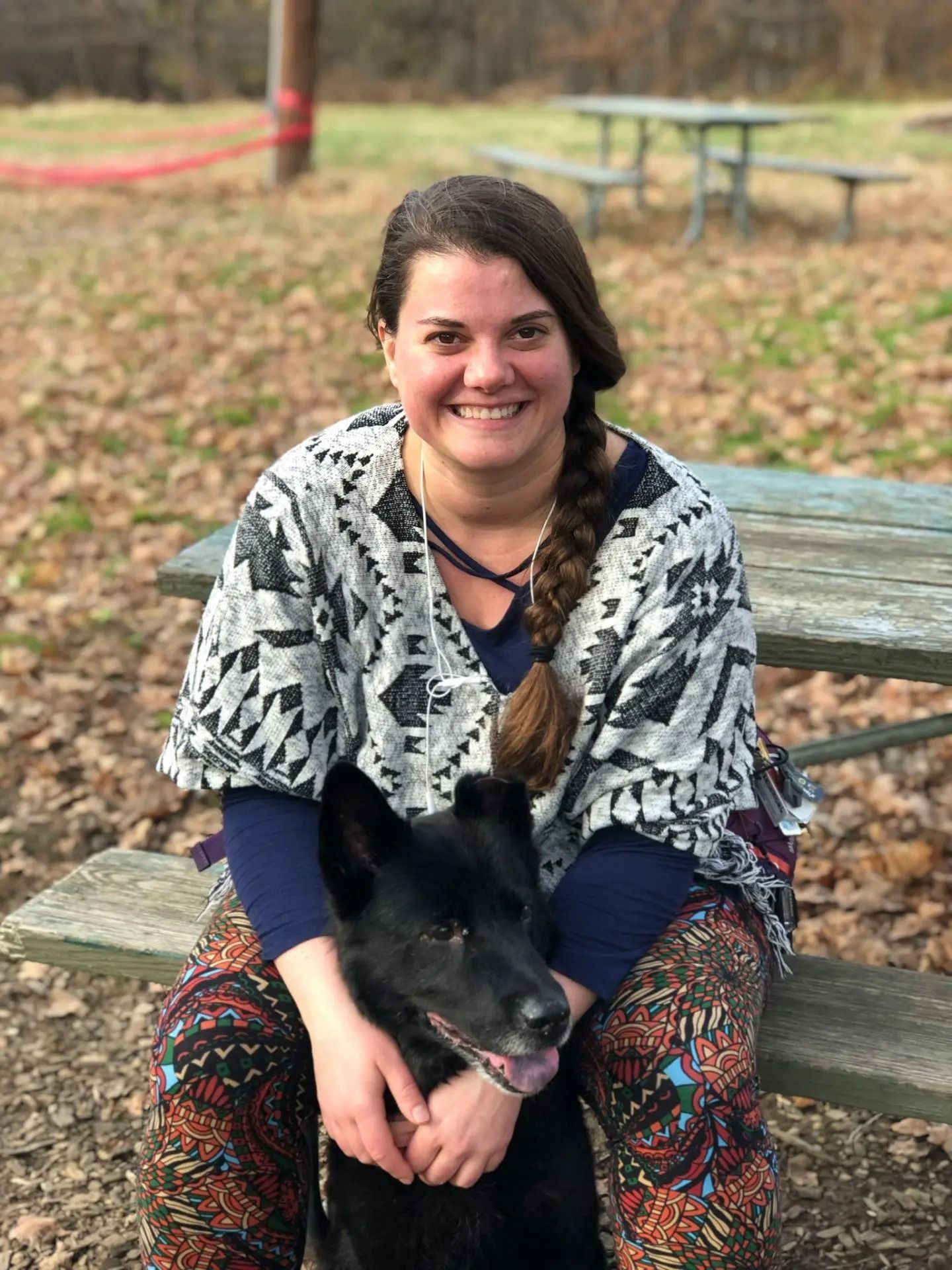 A person smiling while sitting on a picnic table and holding a black dog in an outdoor setting with dry leaves.