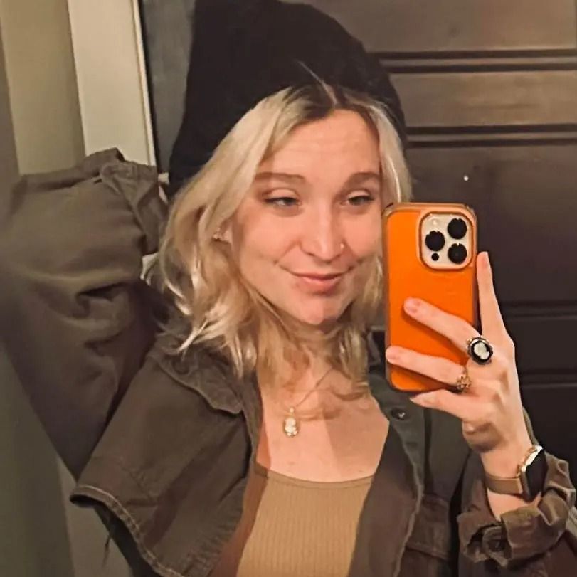A blonde person in a beanie and olive jacket takes a selfie in a mirror, holding an orange-cased phone.
