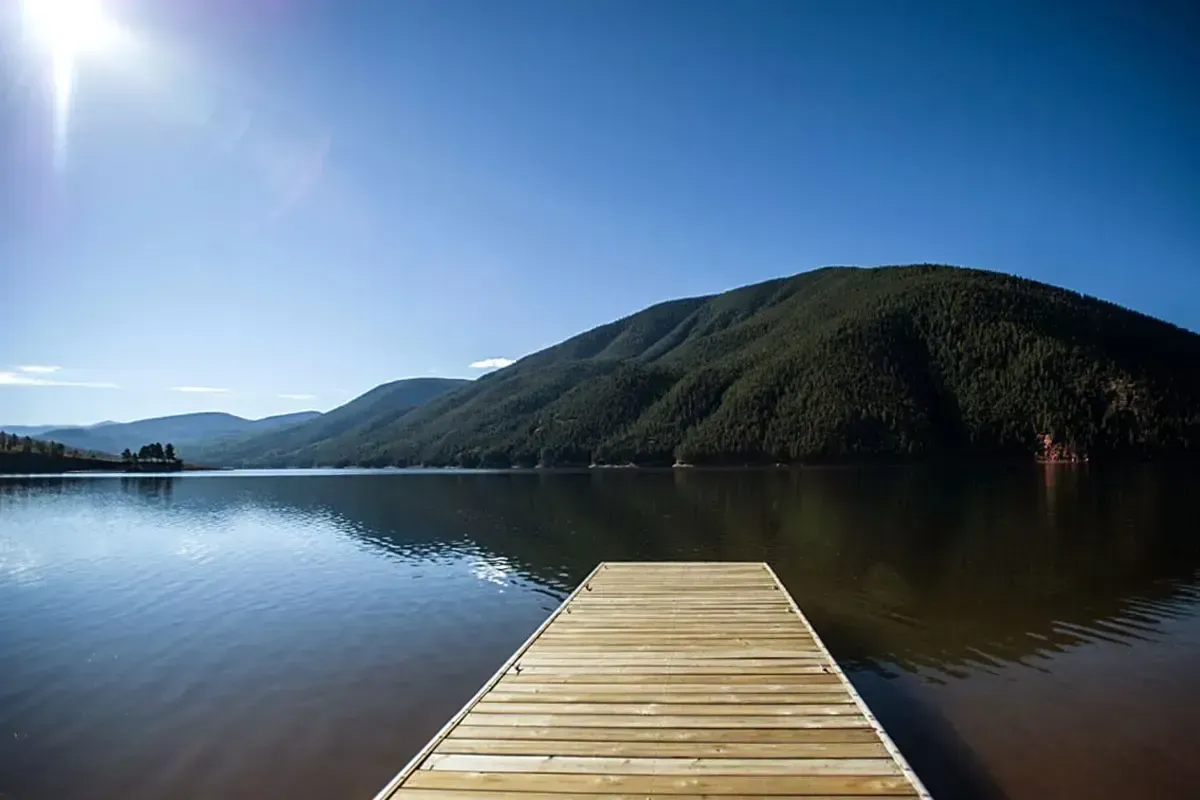 A wooden dock extends into a calm lake, centered before a large, green, sunlit mountain under a clear blue sky.