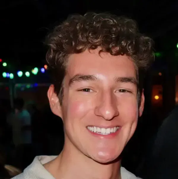 A close-up portrait of a smiling person with curly brown hair against a dark background with blurred, colorful lights.