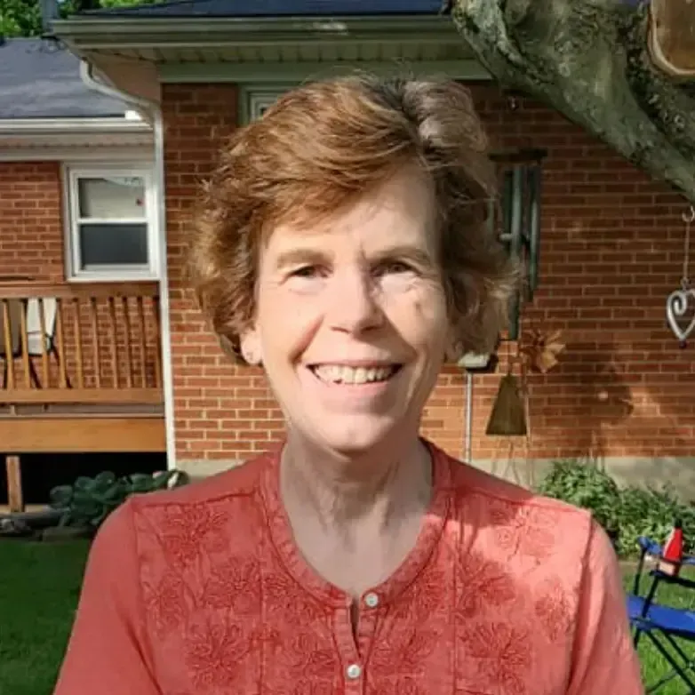 A smiling person with shoulder-length auburn hair wears an orange shirt outdoors in front of a brick house.
