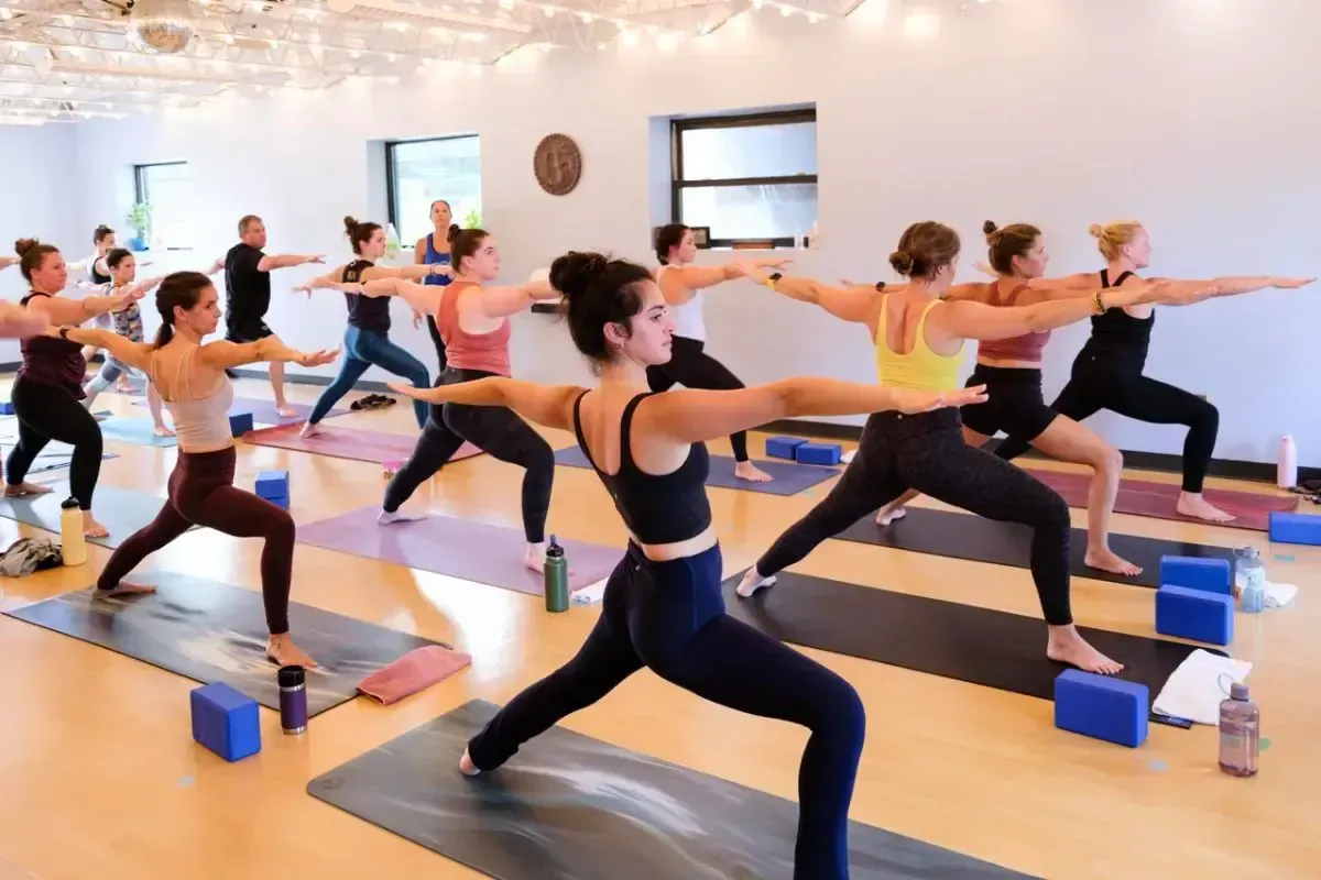 People in a brightly lit studio practice the Warrior II yoga pose on mats, with yoga blocks placed on the floor nearby.