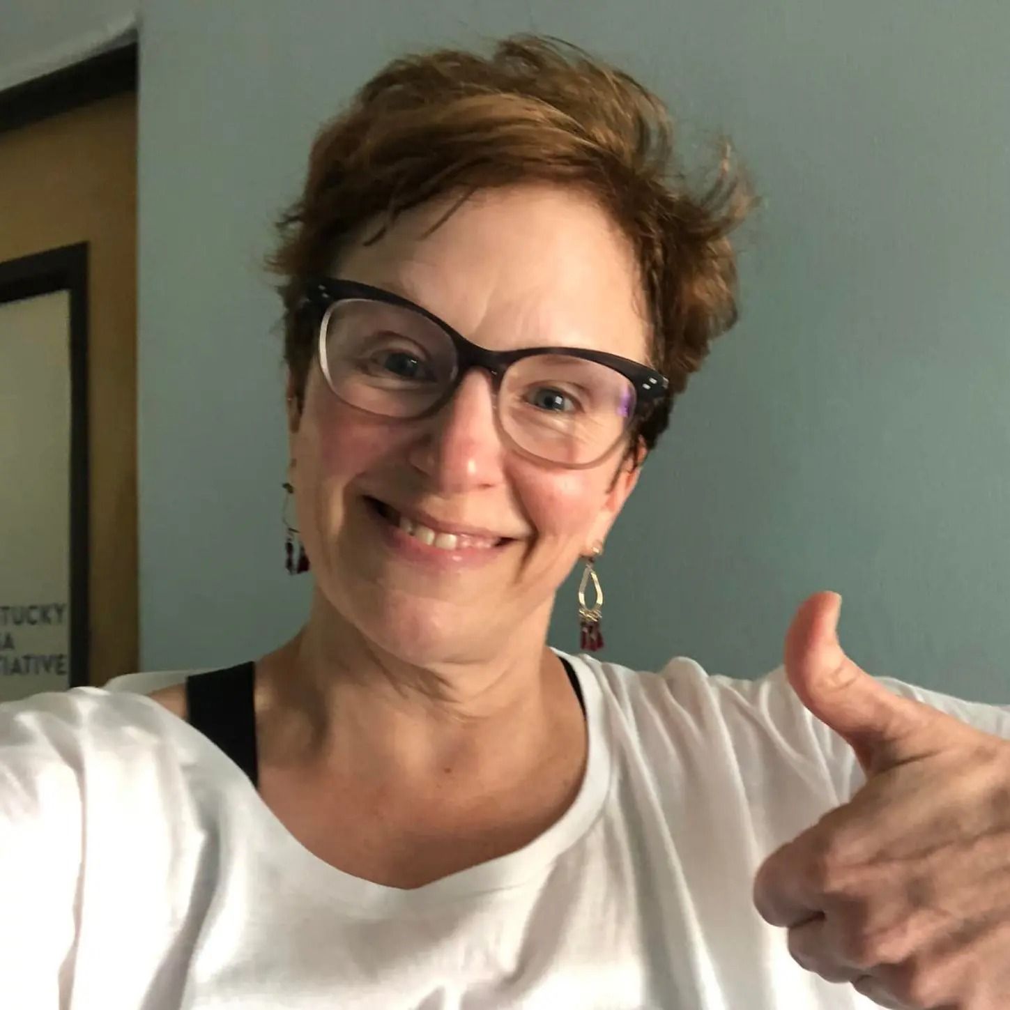 A person with short, reddish-brown hair and glasses smiling and giving a thumbs up in front of a blue wall.