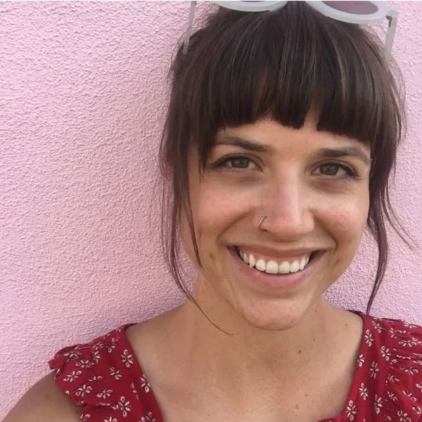 A smiling person with dark bangs and a nose ring, wearing a red patterned top against a textured pink background.