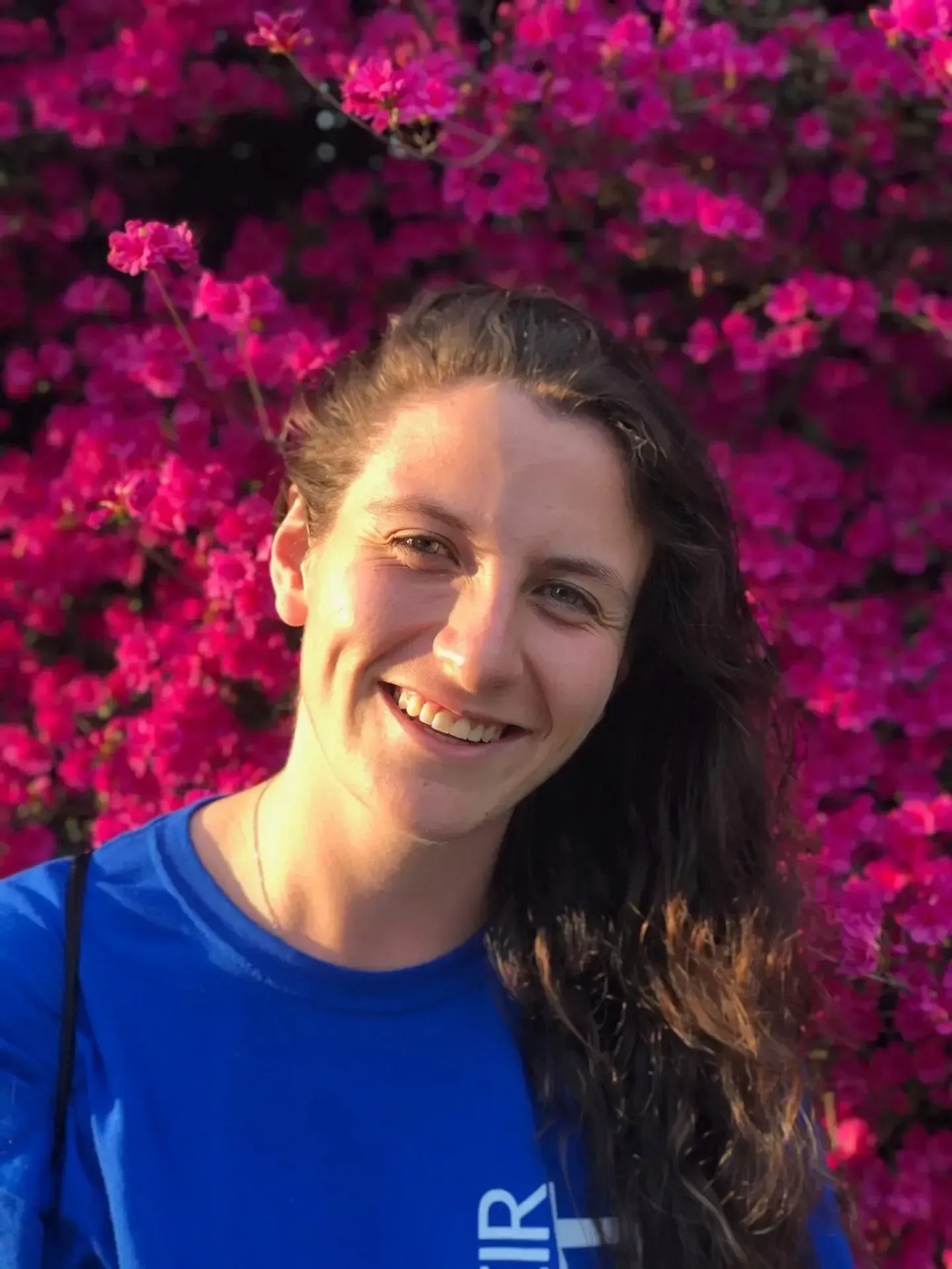 Smiling person with long, dark, wavy hair, wearing a blue shirt against a background of vibrant pink flowers.