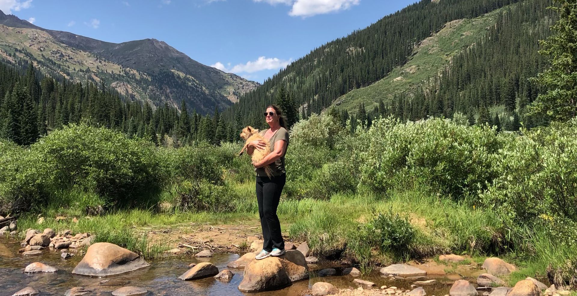 Person standing on rocks by a mountain stream, with green forested hills and blue sky in the background