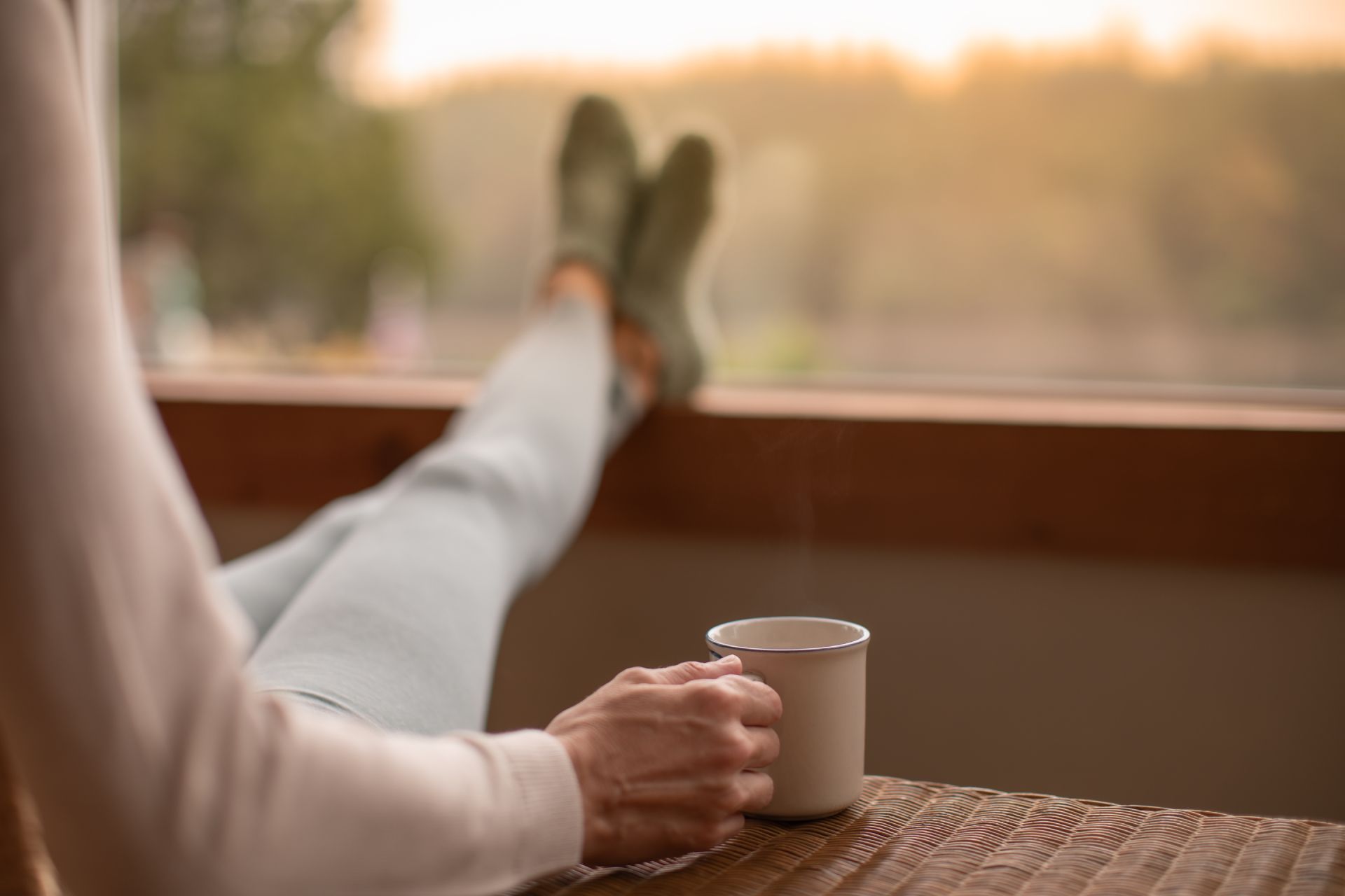 Person relaxing by a window, holding a mug with legs crossed on a woven chair
