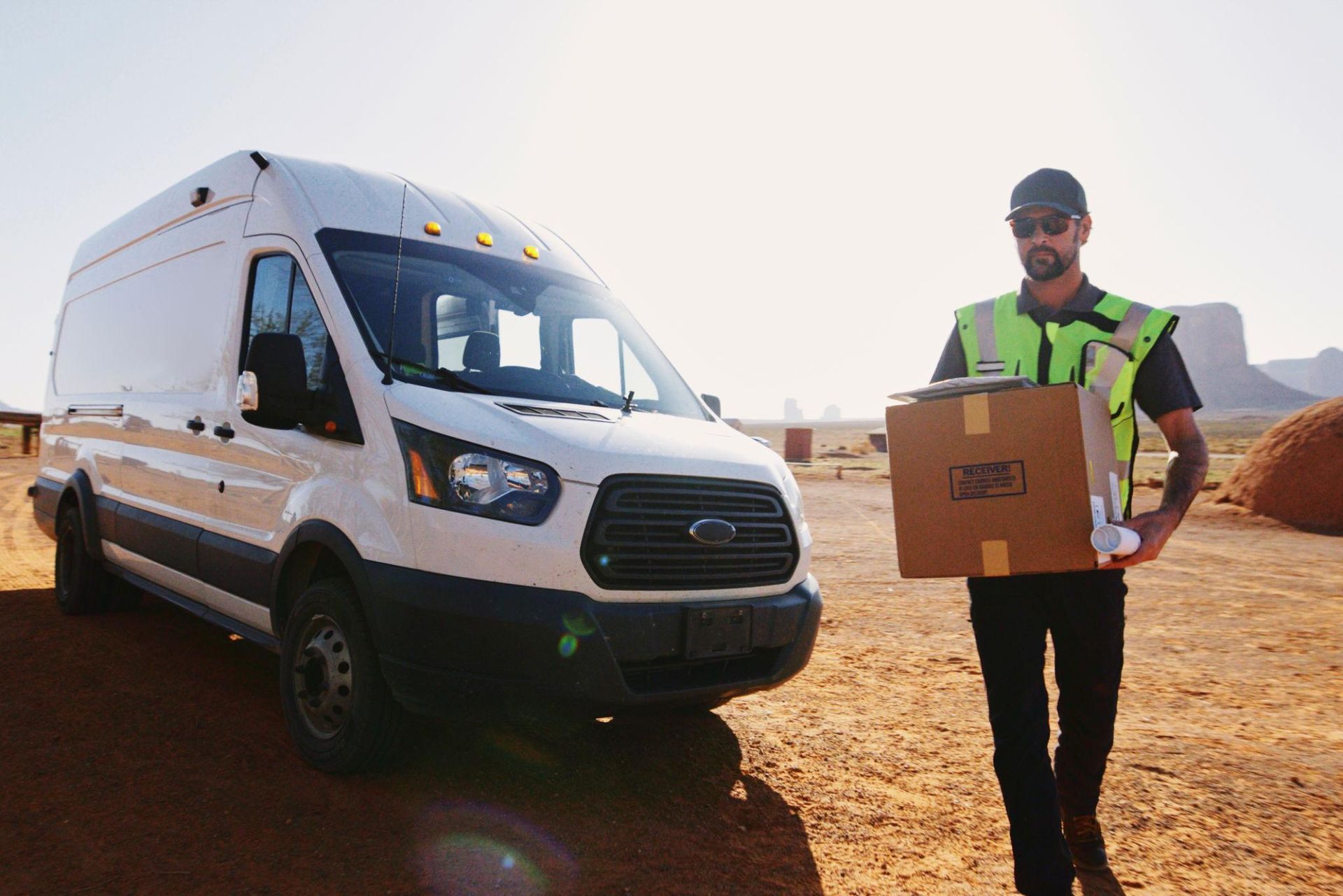 Delivery worker in neon vest carrying a box beside a white cargo van on a dirt road at sunset