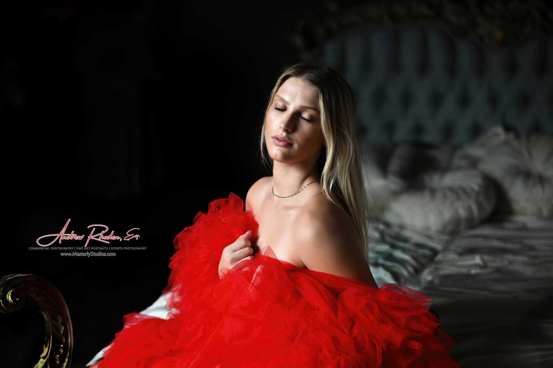 Woman in a red tulle gown posing on a bed in a dimly lit bedroom