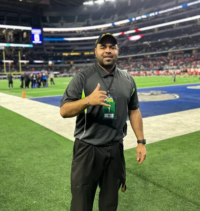 Man in a black and grey uniform gives a thumbs-up on a football field. He wears a hat and lanyard. Stadium in background.