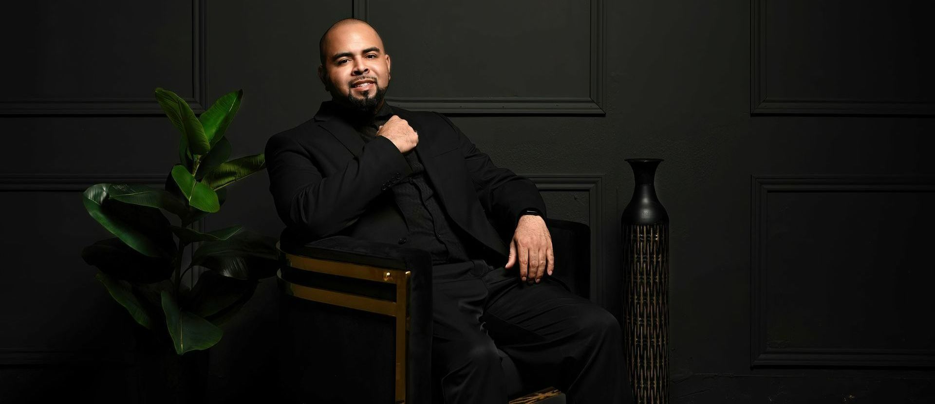 Man seated in a dark studio chair beside a leafy plant, wearing black against a black paneled wall.
