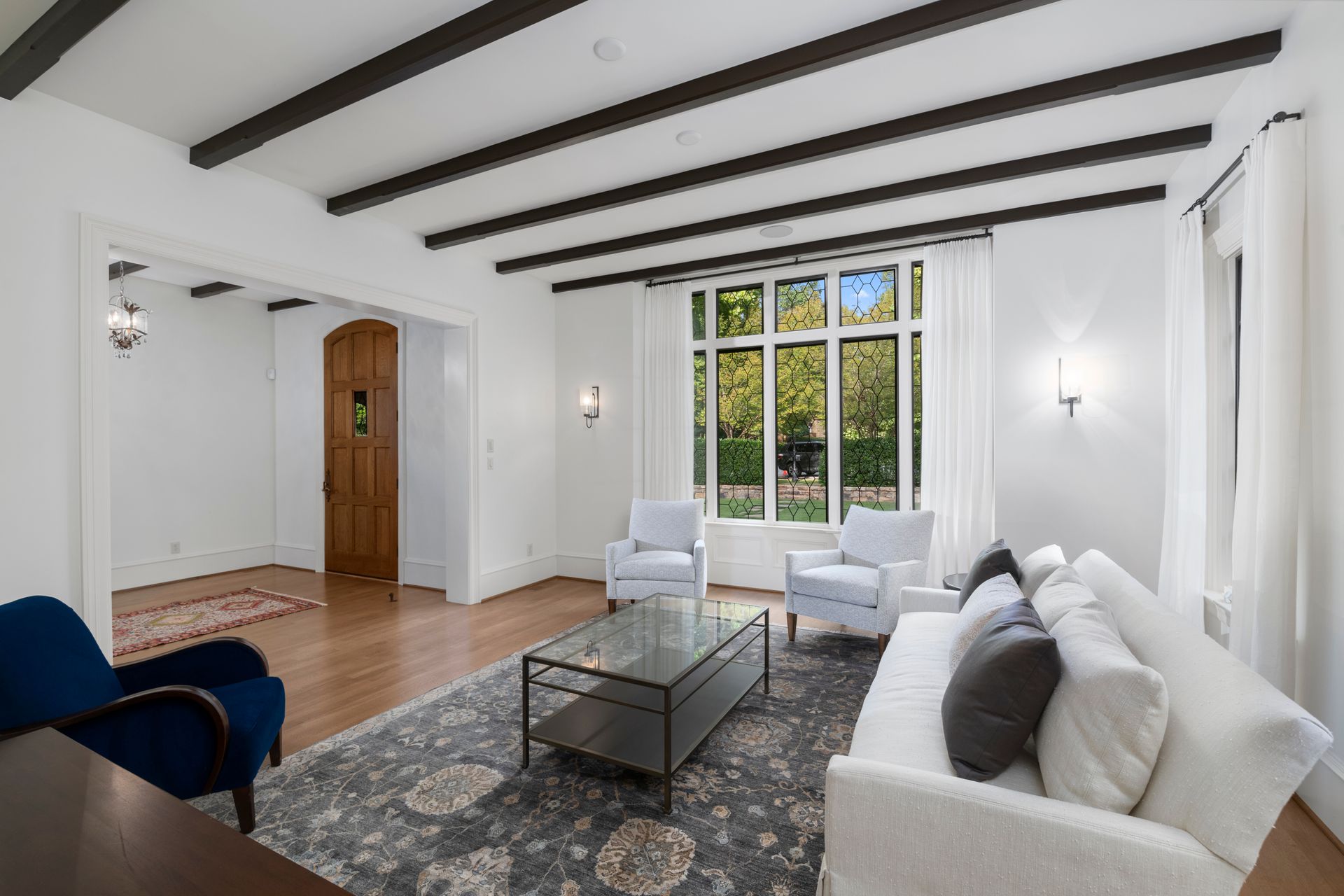 Bright living room with white walls, exposed beams, a sofa, chairs, and large windows overlooking trees