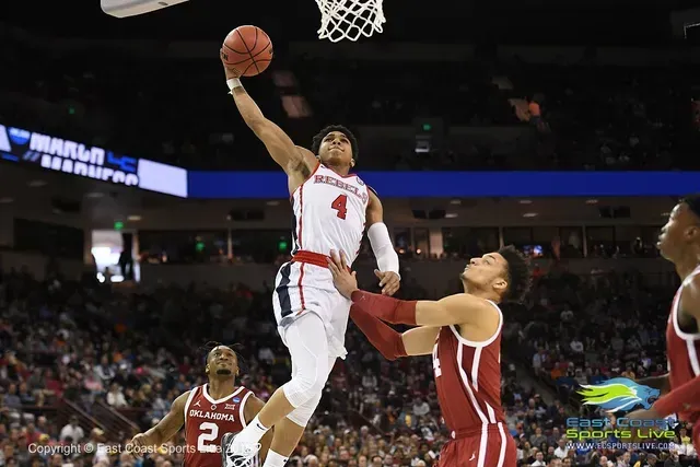 Basketball player in a red jersey shoots a layup, defender attempts block. Indoor court, crowd in background.