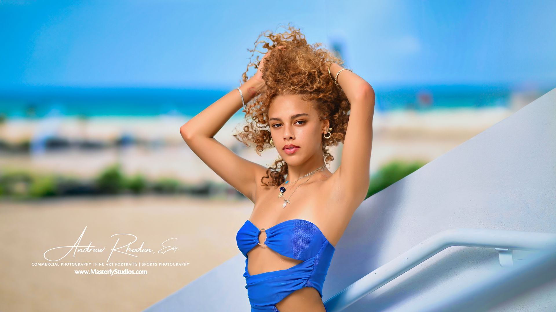 Woman in a blue swimsuit posing on a beach with hands in her hair, ocean blurred behind her