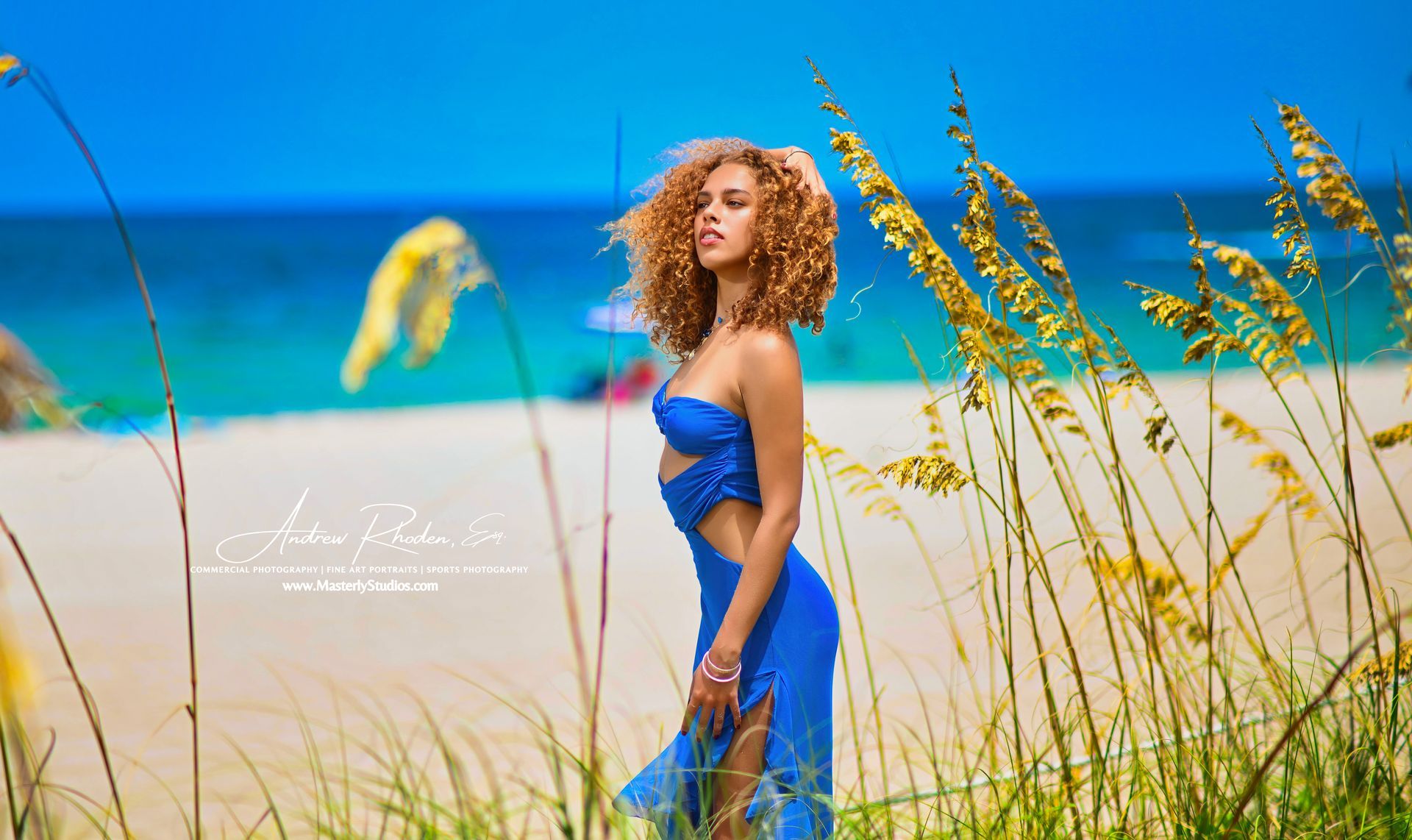 Woman in a blue dress standing in tall grass on a sandy beach under a bright blue sky