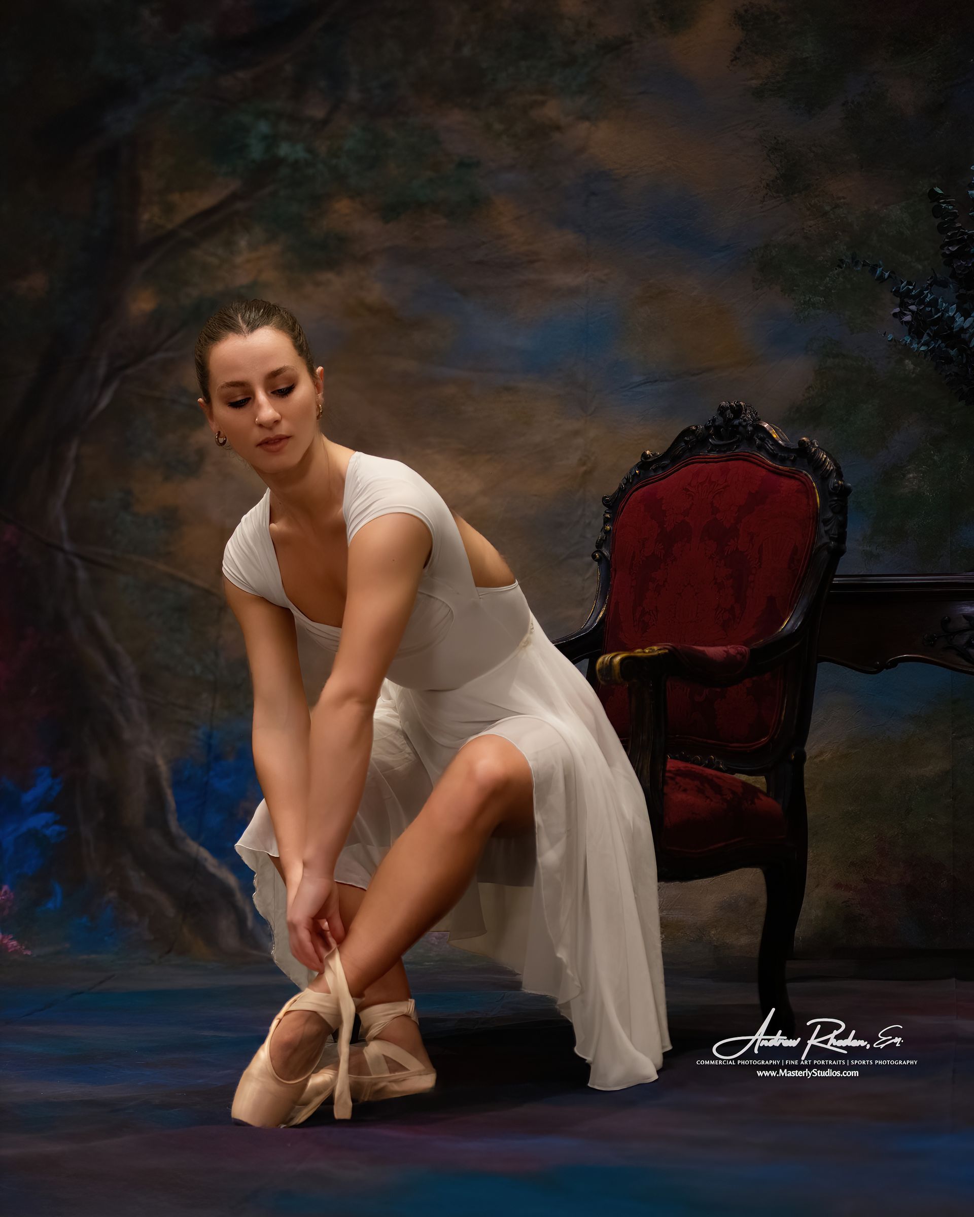 Ballet dancer in a white dress adjusting pointe shoe beside a red velvet chair on a dark stage backdrop