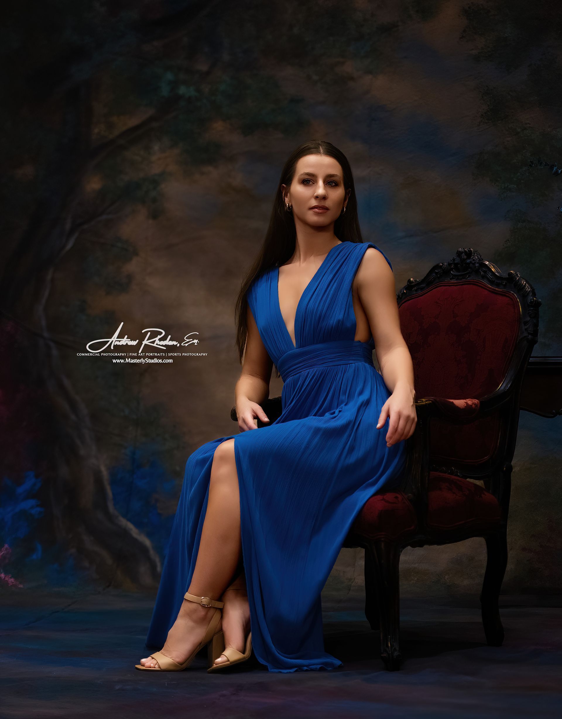 Woman in a blue dress seated on a dark red chair against a moody studio backdrop