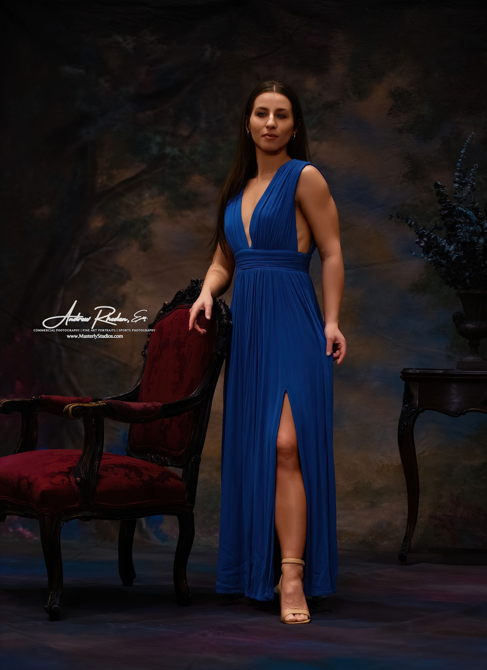 Woman in a blue evening gown standing beside a red velvet chair in a dark studio setting