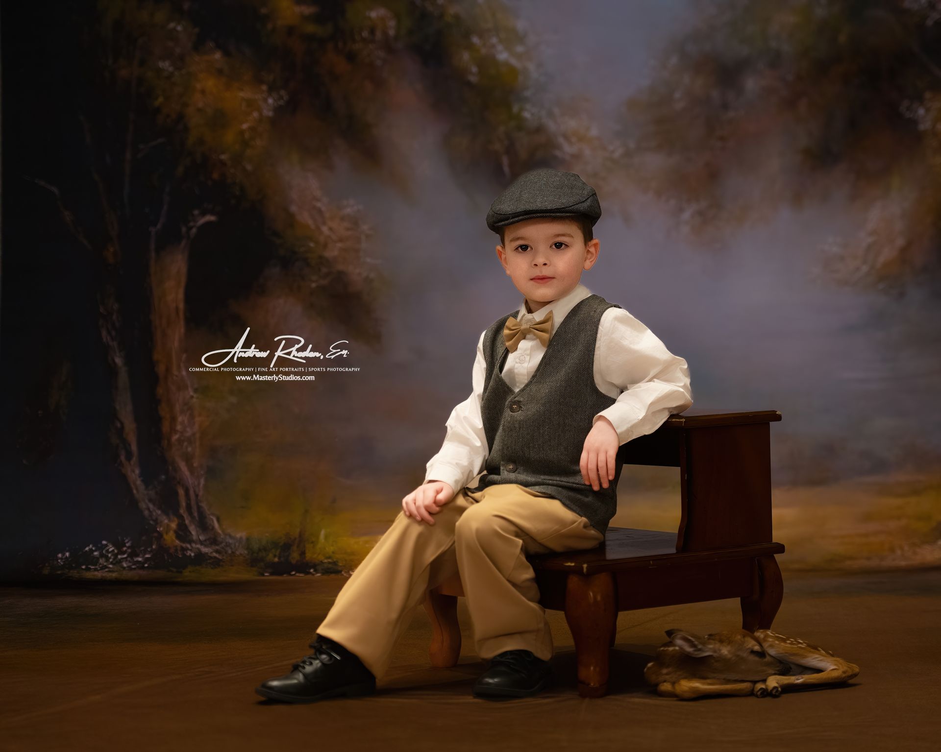 Child in cap and vest seated on a wooden bench in a studio portrait, with a painted forest backdrop.