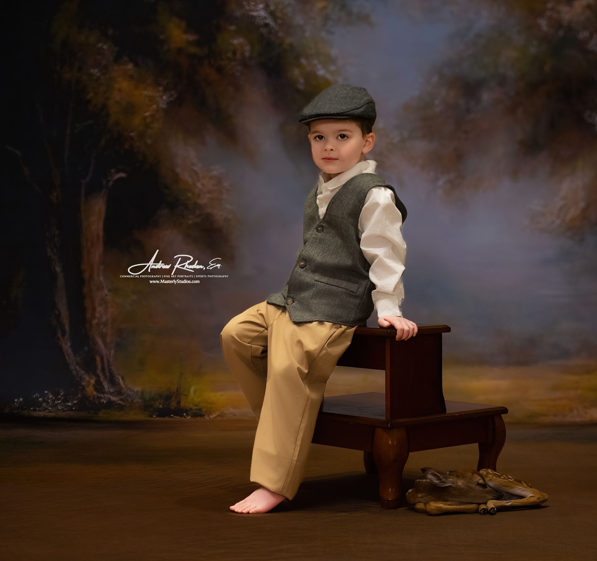Child in a flat cap and vest sits on a wooden stool in a studio portrait, bare feet on floor.