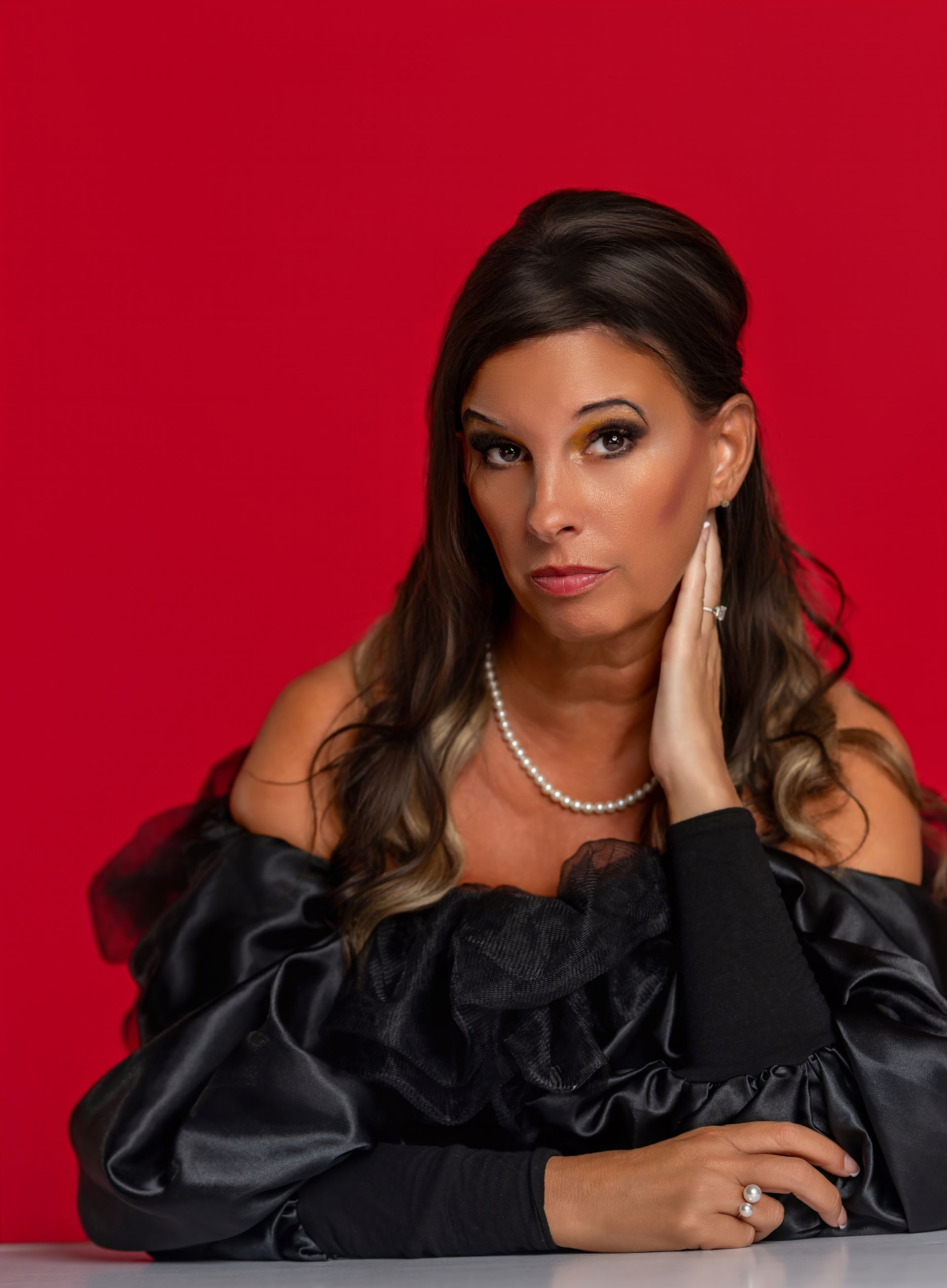 Portrait of a woman in a black off-shoulder dress against a red background, resting her face on her hand