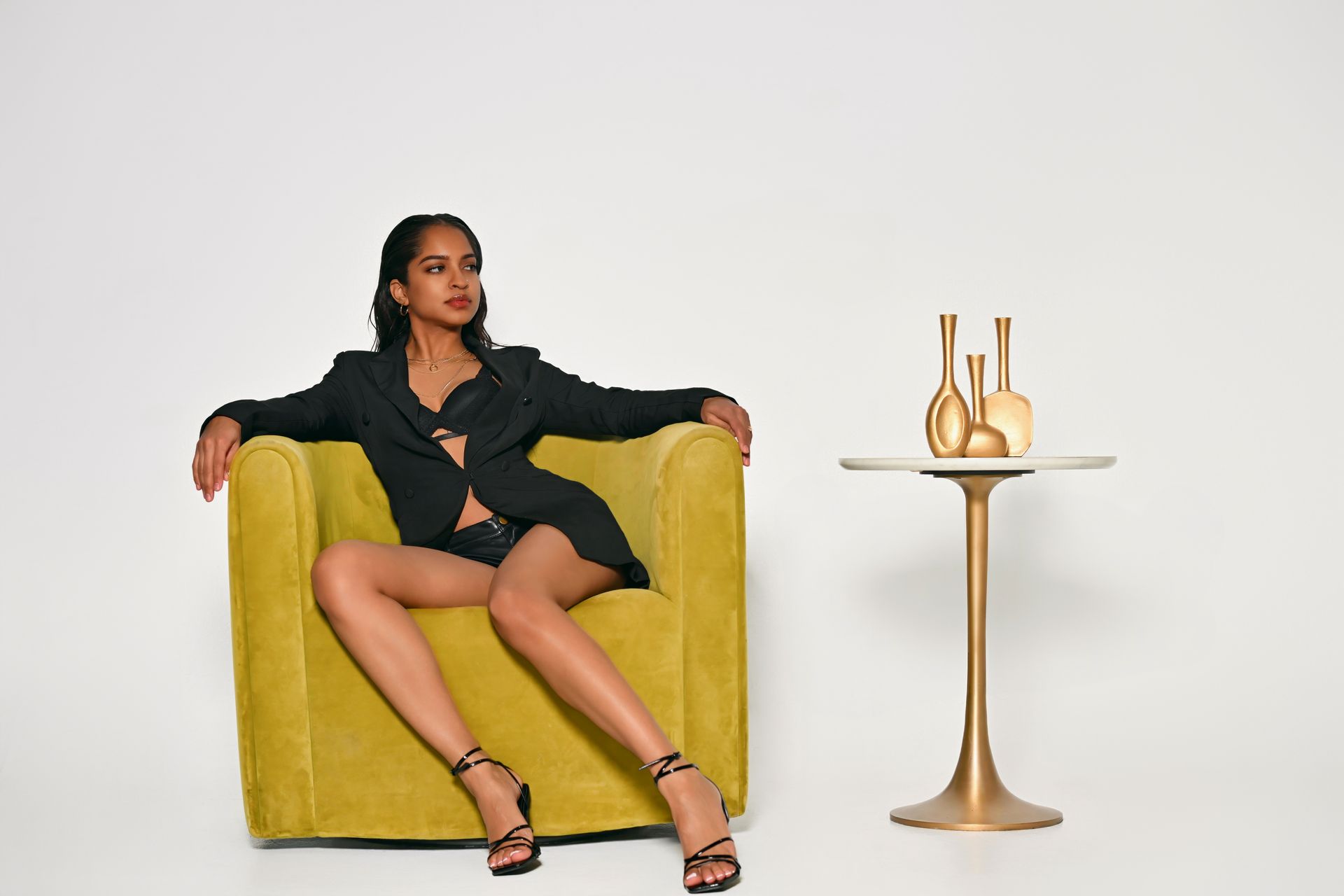 Woman seated on a yellow armchair beside a small gold side table with vases on a white background