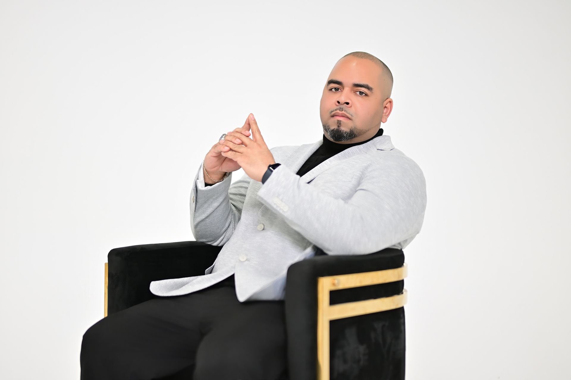 Man in a white blazer seated in a black chair, holding his hands together against a plain white background