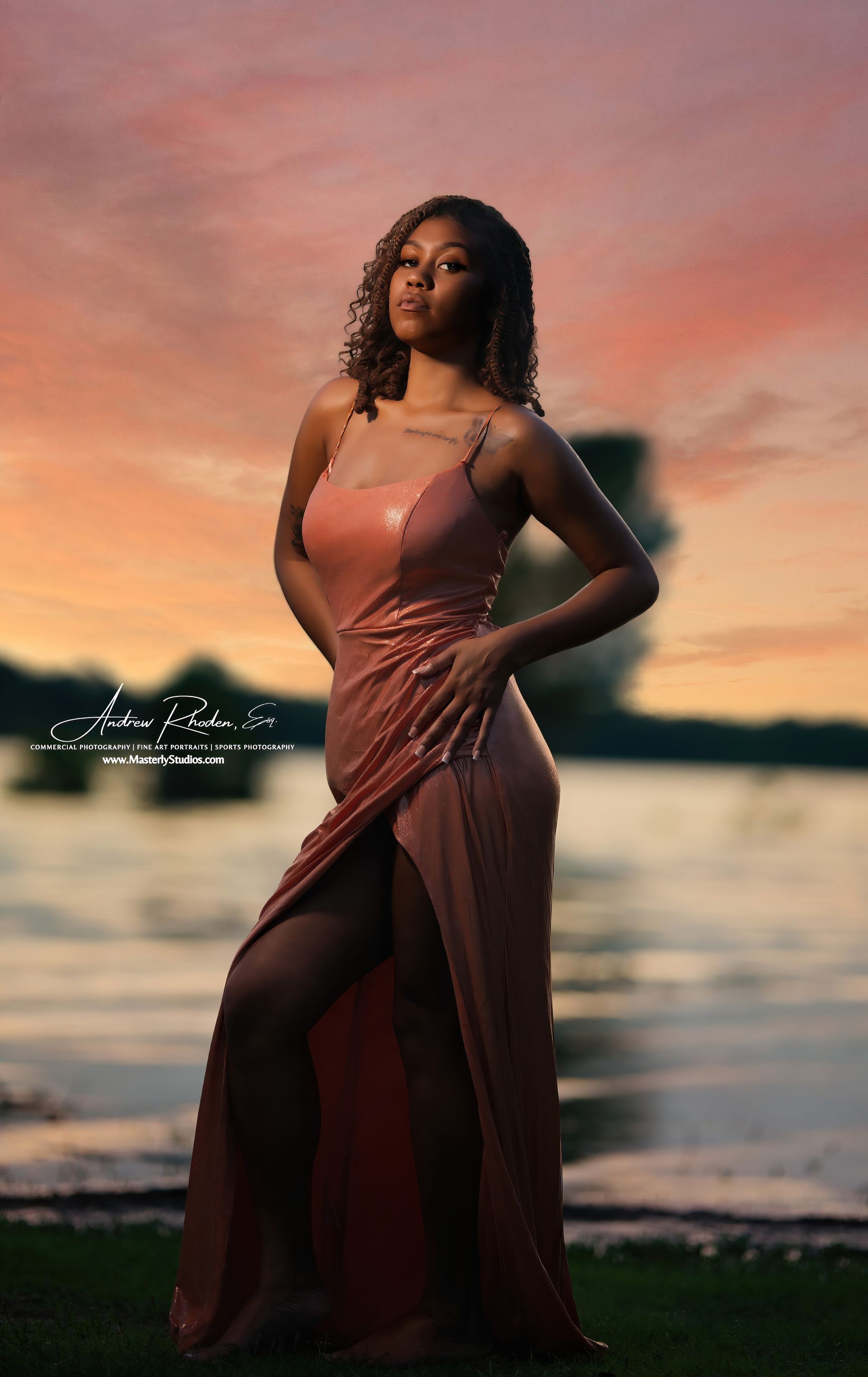 Woman in a rust-colored gown standing by the beach at sunset, with water and a pink-orange sky behind her.